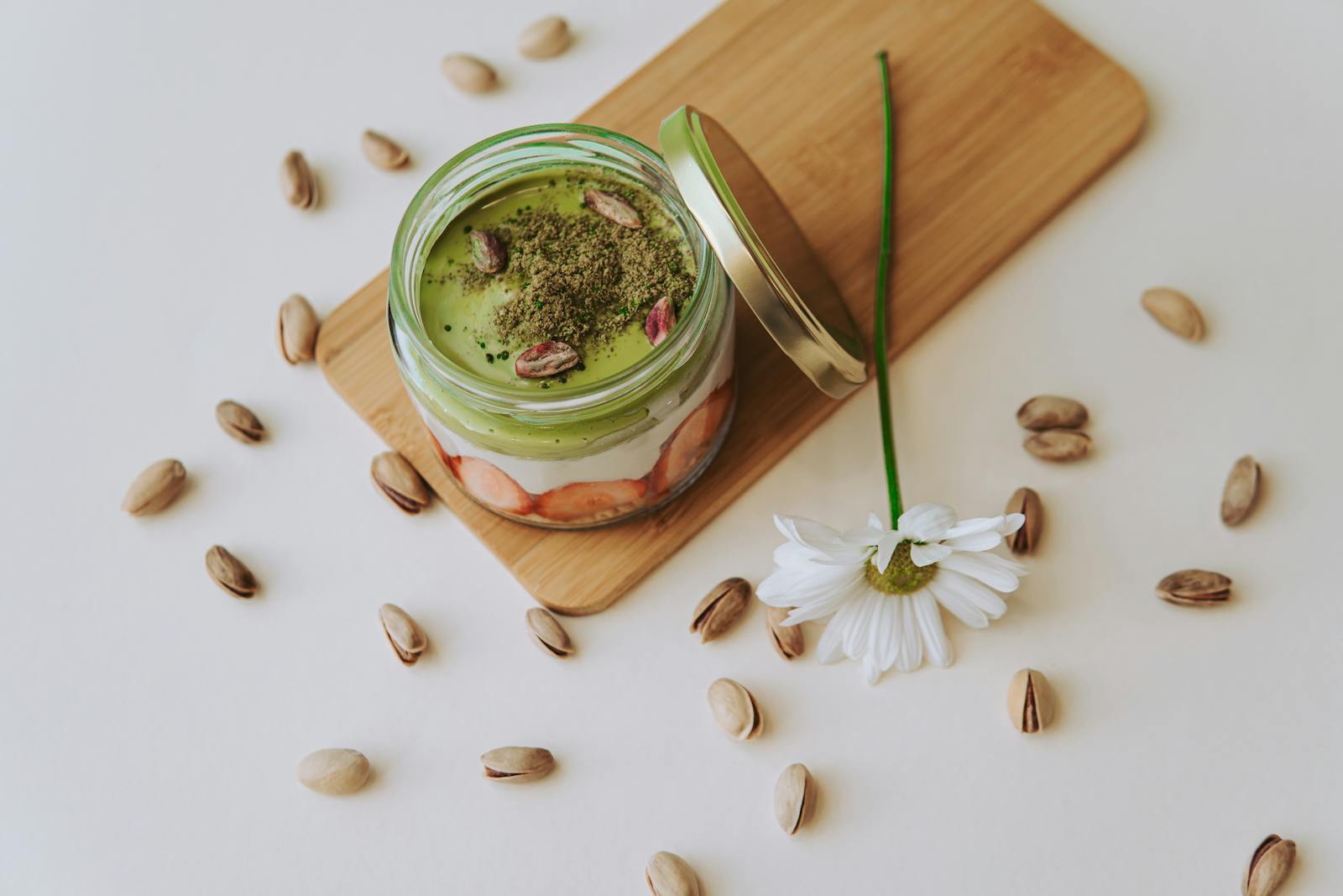 A stylish layered dessert jar with matcha, berries, topped with pistachios, accompanied by a flower, on a wooden board.